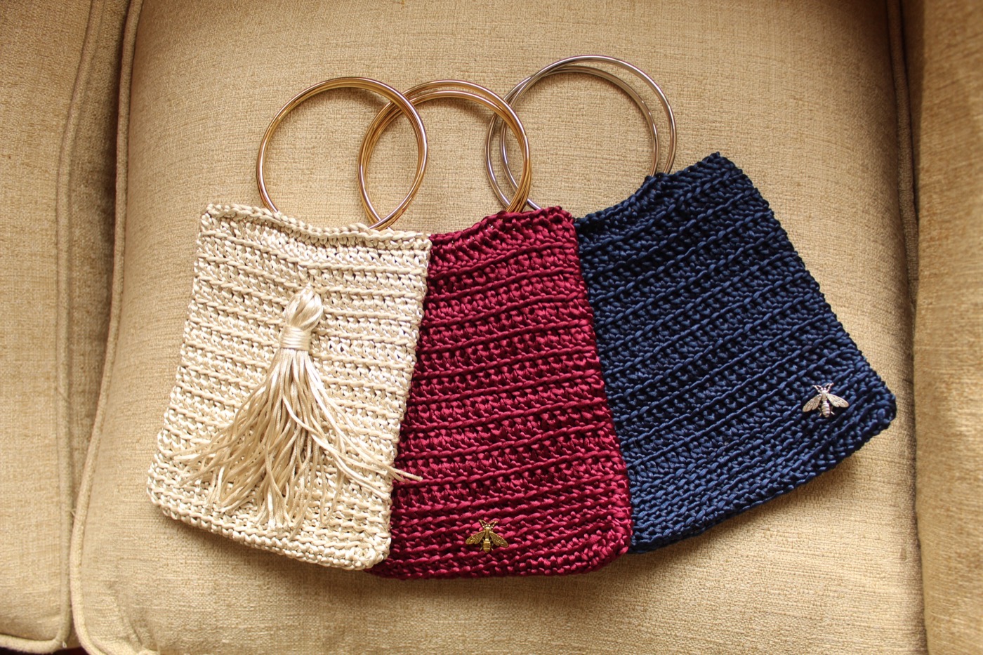 Tres carteras de crochet en crudo, burdeo y azul, con asas doradas